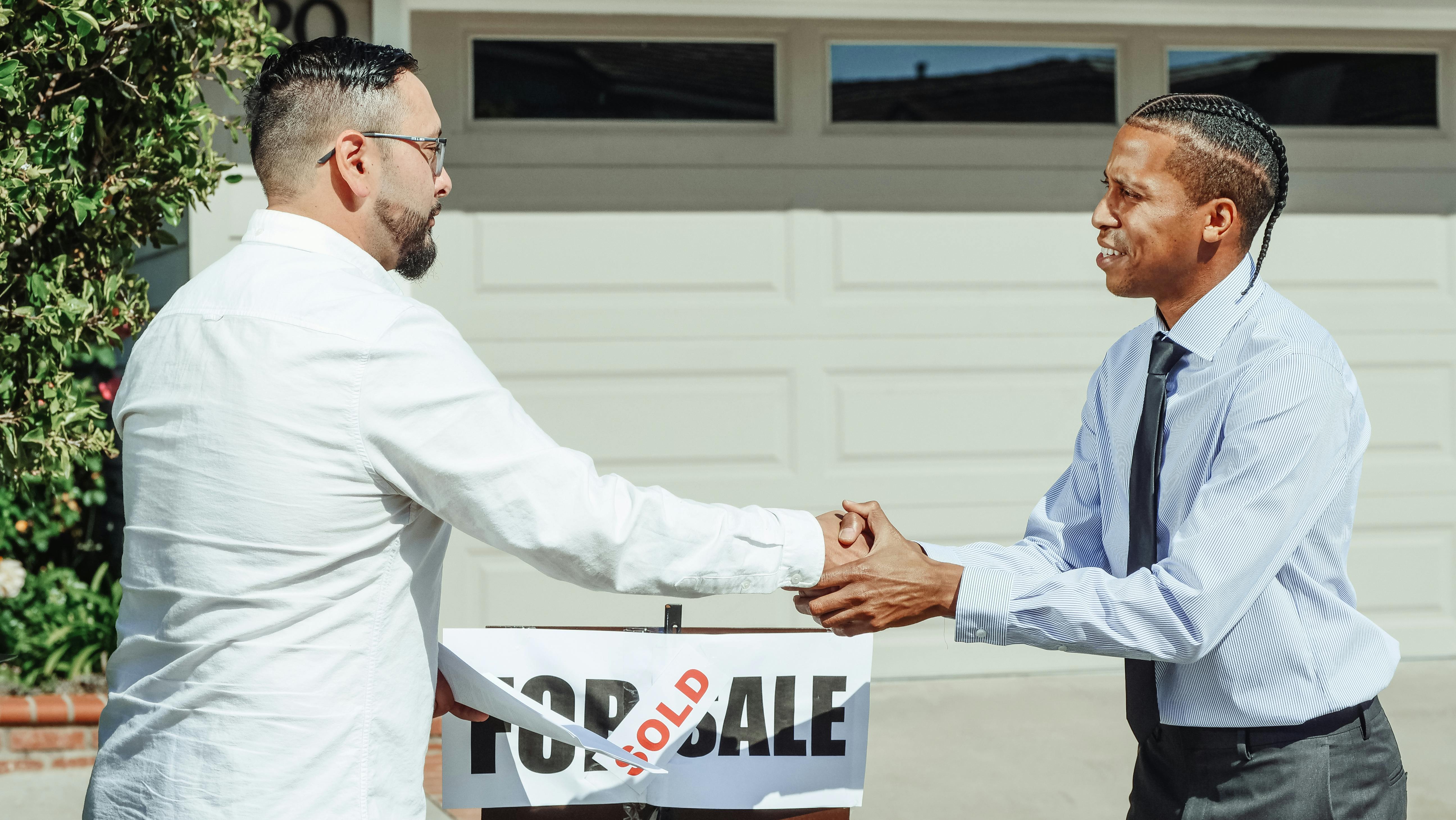 Two men shaking hands in front of a sold house sign